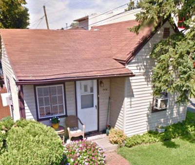Small white house with a brown roof and a front porch with chairs.