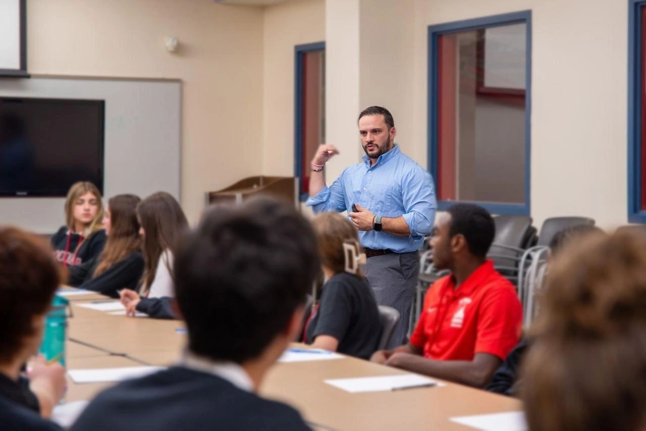 A man in a blue shirt leads a discussion with students around a table.