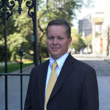 Man in suit and yellow tie standing outdoors near a wrought iron gate.