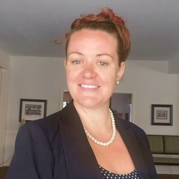 Smiling woman in a black blazer and pearl necklace indoors.