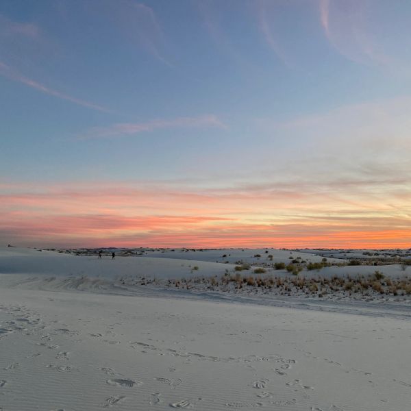 Sunset over white sand dunes with footprints and sparse vegetation.