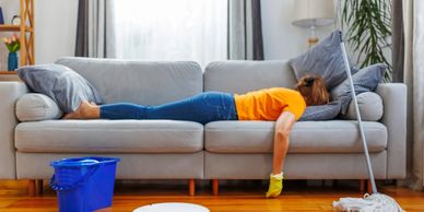 Tired woman lying on couch after cleaning, with mop and cleaning tools nearby.