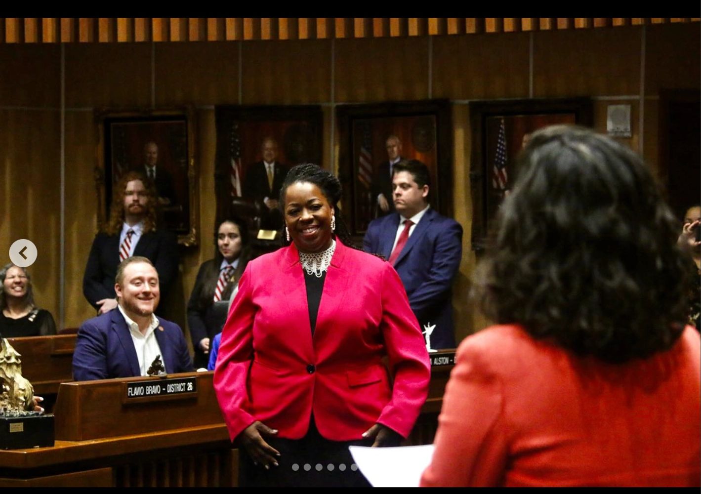 Woman in a pink blazer smiles confidently in a formal meeting room.