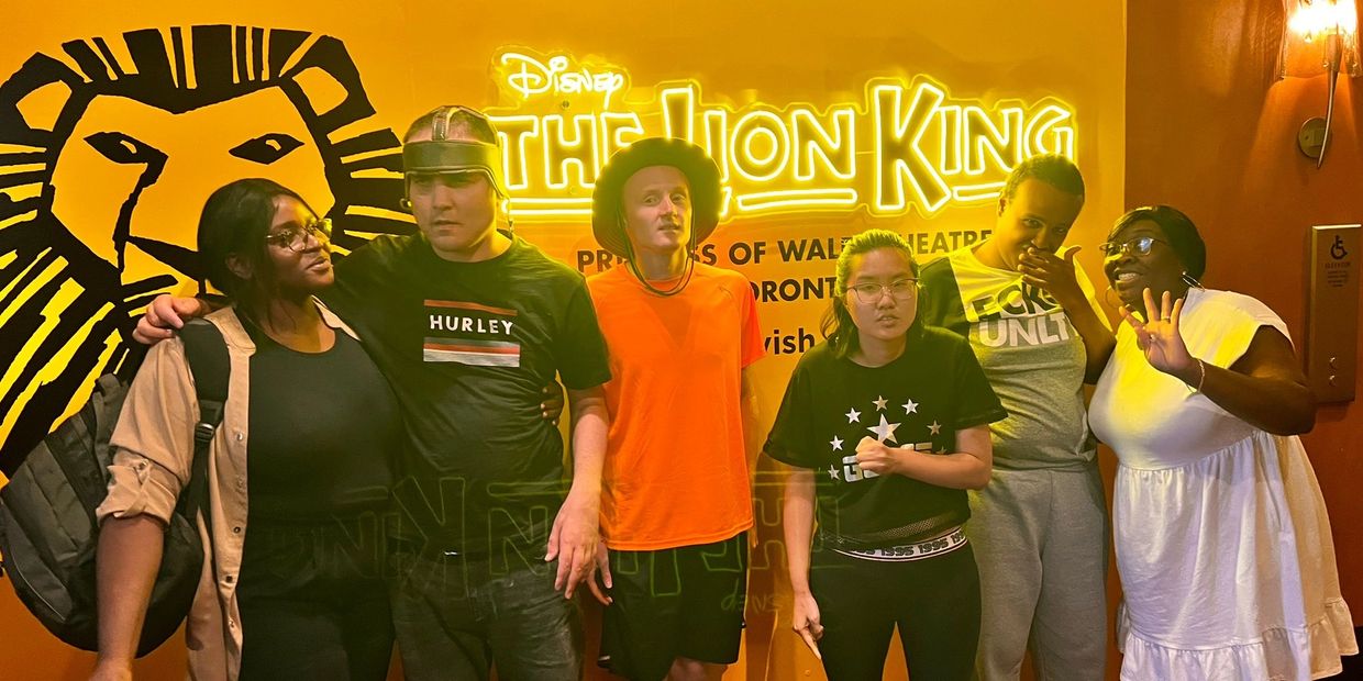 Group of six people posing in front of a bright Lion King sign.