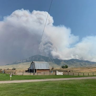 Billowing smoke from a wildfire behind a rustic barn in a rural landscape.