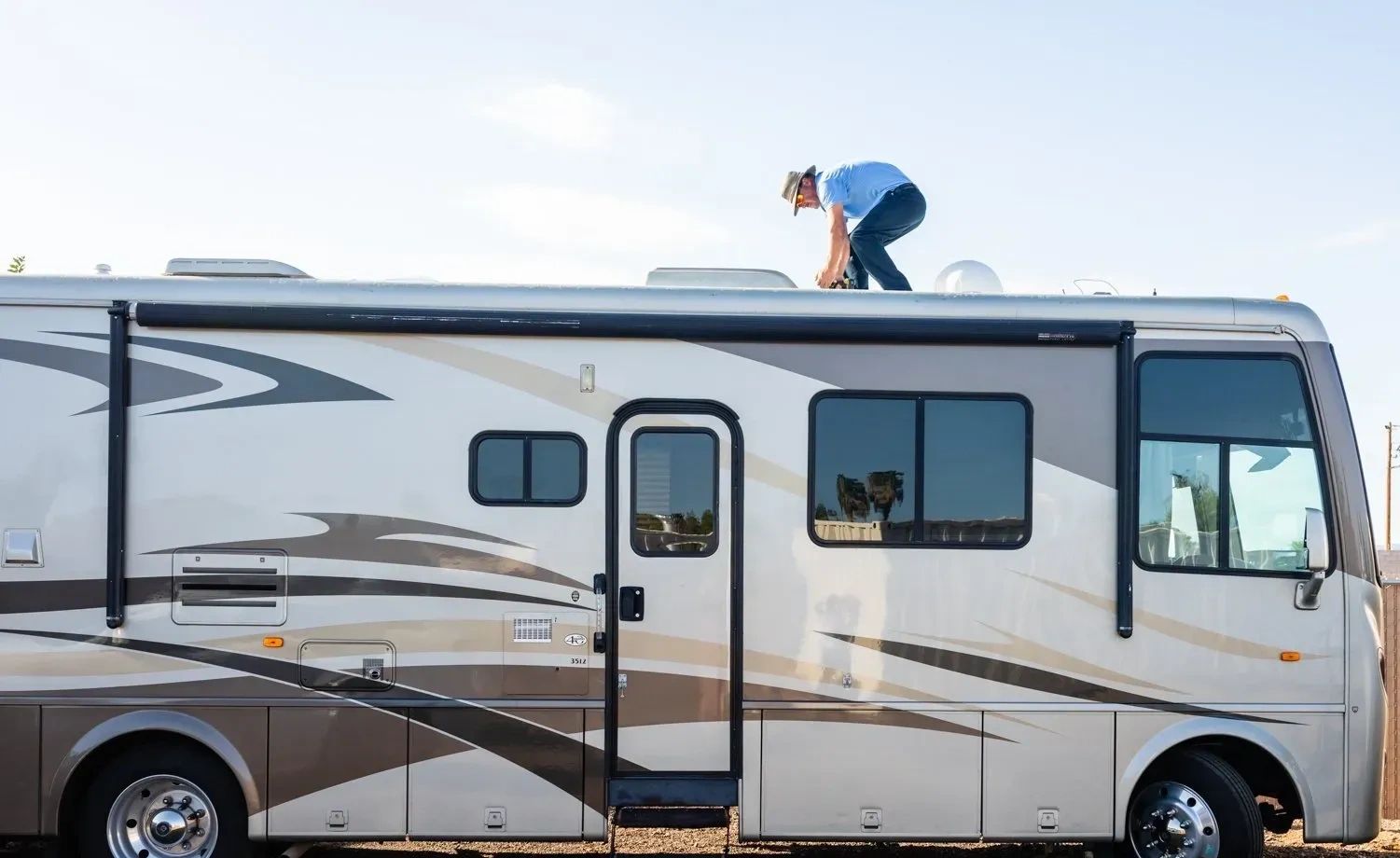 Man in a hat working on top of a recreational vehicle under clear sky.