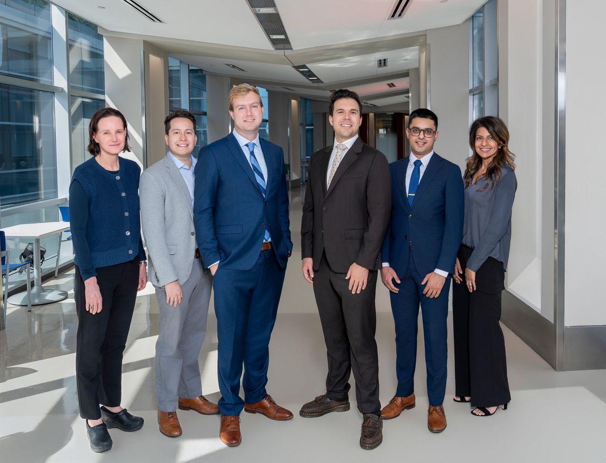 Six professionals posing together in a modern office hallway, dressed in business attire.