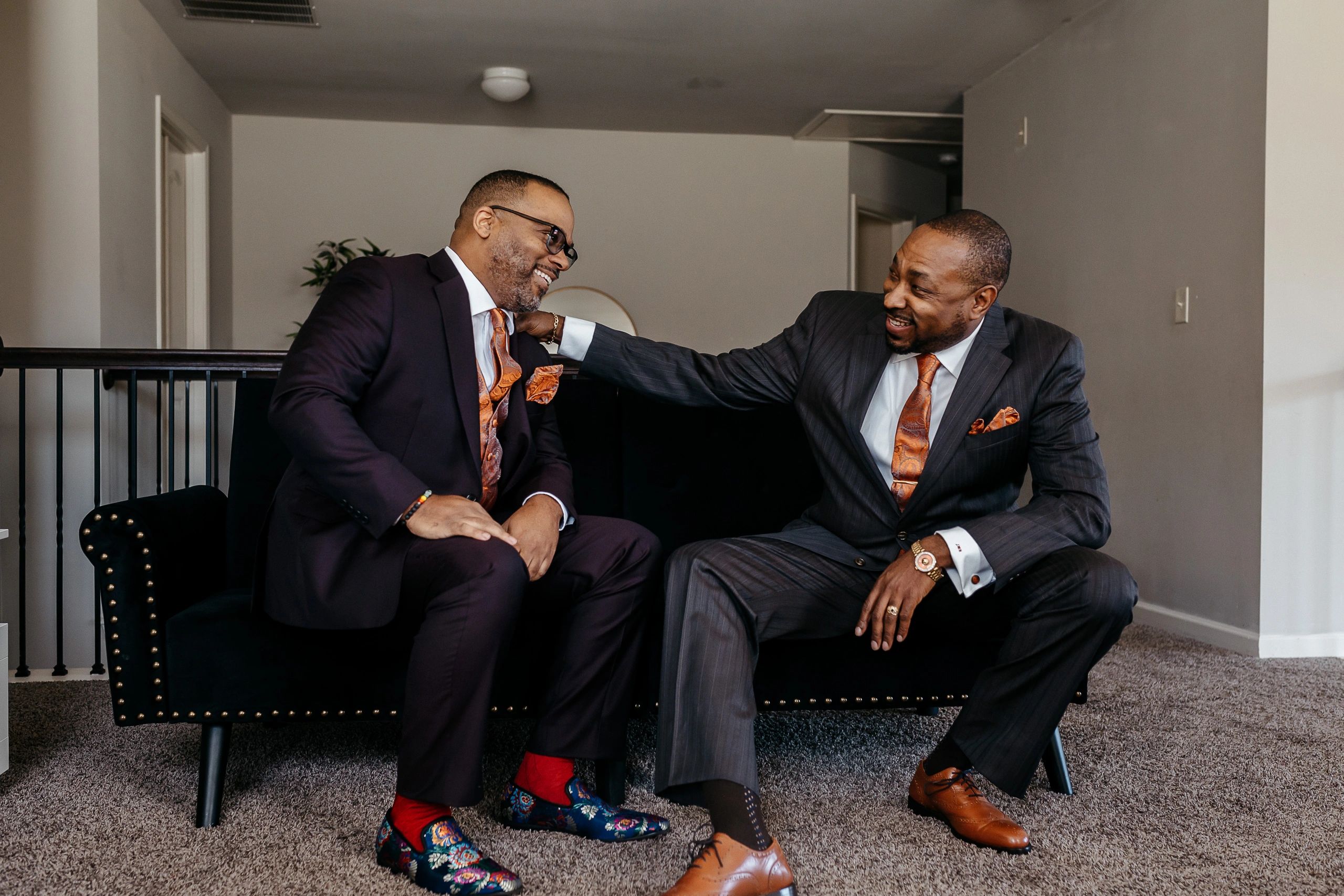 Two men in suits sharing a joyful moment on a black couch indoors.