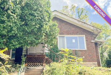 Brick house with a front porch surrounded by greenery and a pending sale sign.
