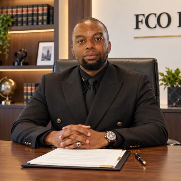 Man in black suit sitting at a desk with documents.