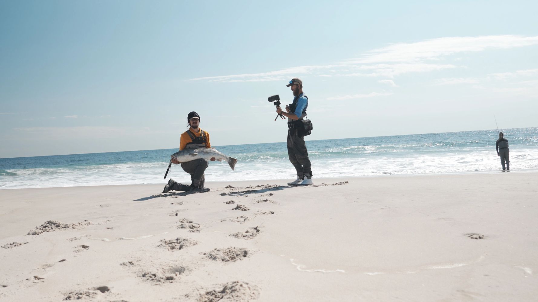 Man kneels on beach holding large fish while another films him near the ocean.