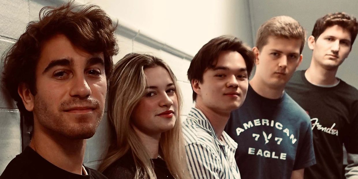 A diverse group of five young adults posing indoors against a wall.