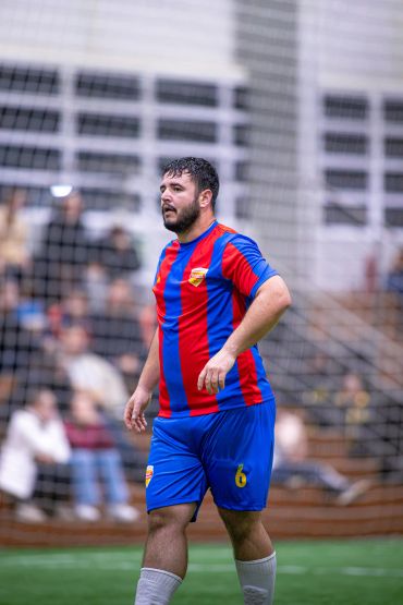 Jogador de futebol com uniforme azul e vermelho em quadra indoor.
