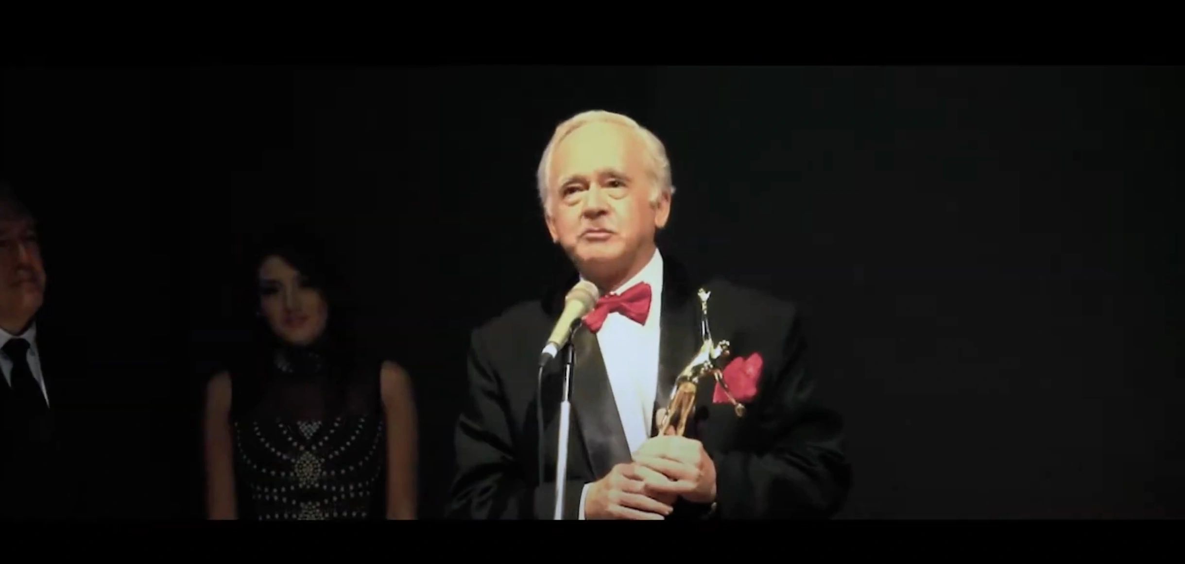A man in a tuxedo holds an award while speaking at a microphone on stage.