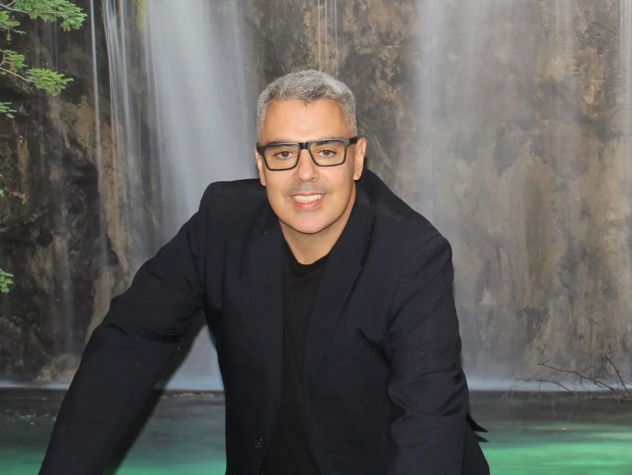 Homem usando um blazer na cor escura, com uma imagem de cachoeira aparecendo ao fundo