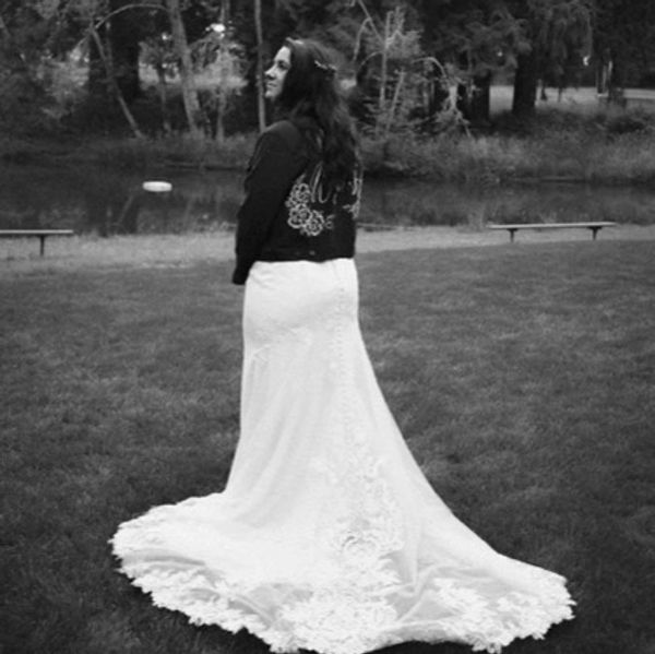 Bride in a white wedding dress with a black jacket, standing outdoors on grass.