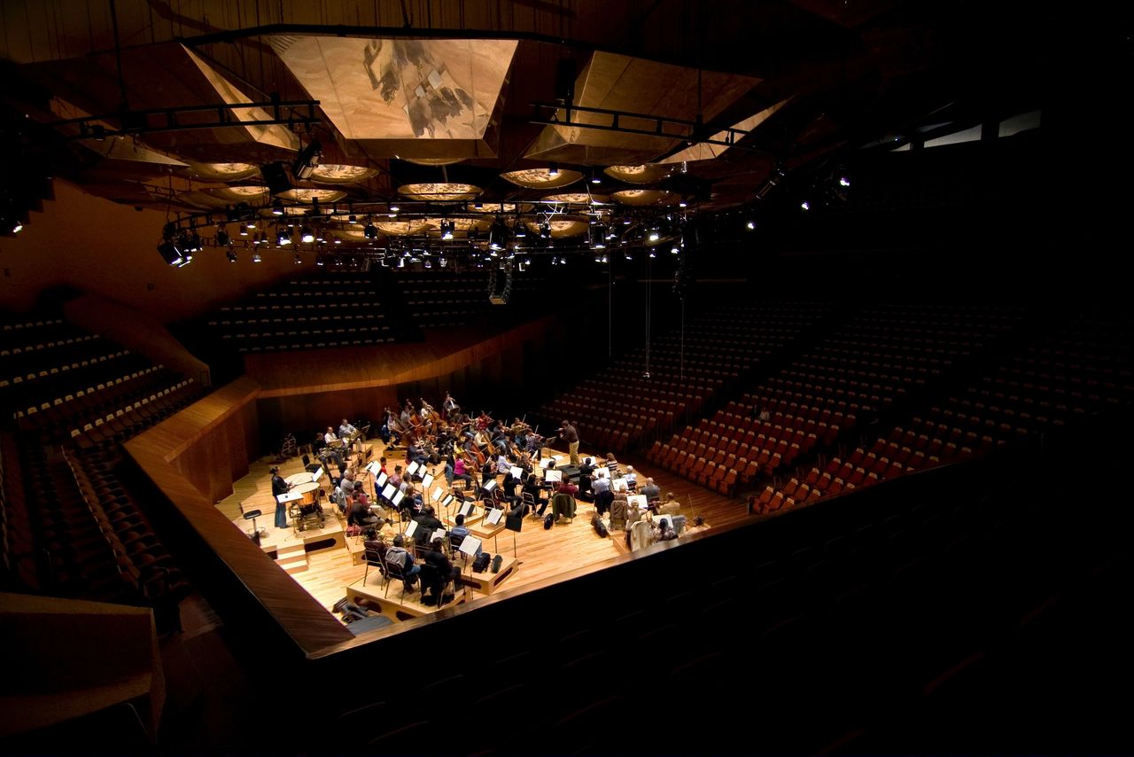 This image captures the serene atmosphere of an empty symphony hall during a rehearsal, showcasing an orchestra in mid-practice with a conductor at the helm. The grandeur of the concert hall, with its rows of vacant seats, spotlights focused on the stage, and the warm hues of the wooden interior, suggests a quiet moment before the musicians share their artistry with an audience. The image conveys a sense of anticipation and the disciplined art of musical performance. This image captures the serene atmosphere of an empty symphony hall during a rehearsal, showcasing an orchestra in mid-practice with a conductor at the helm. The grandeur of the concert hall, with its rows of vacant seats, spotlights focused on the stage, and the warm hues of the wooden interior, suggests a quiet moment before the musicians share their artistry with an audience. The image conveys a sense of anticipation and the disciplined art of musical performance.