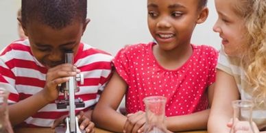 Un niño examina una muestra bajo un microscopio mientras relata lo que ve a dos niñas