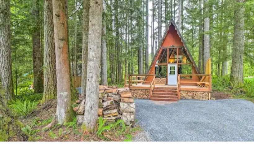 A-frame cabin in forest setting with large front deck, firewood stored underneath, and gravel drive.