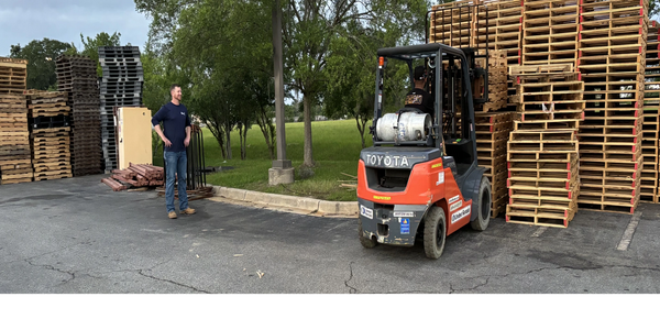 A man operates a forklift stacking wooden pallets outdoors while another man watches.