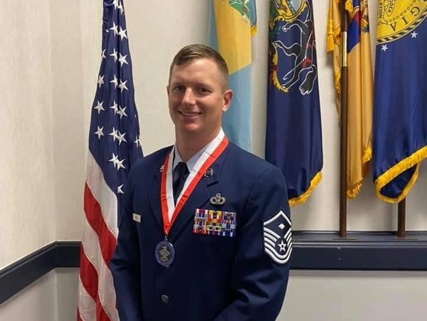A smiling military officer in dress uniform with medals, standing by flags indoors.
