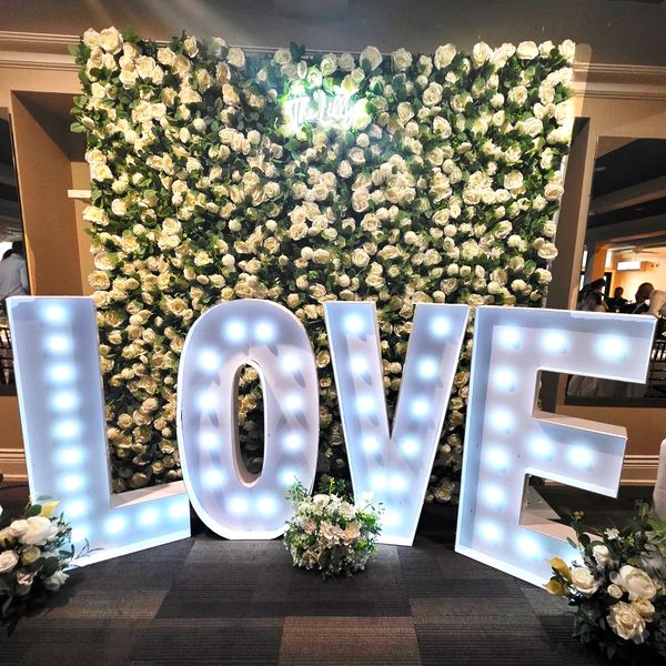 Illuminated LOVE letters in front of a white rose floral backdrop.