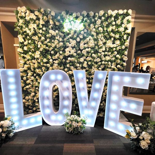 Illuminated LOVE letters in front of a white rose floral backdrop.