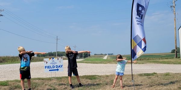 Three children dabbing near a Farmers Field Day sign and flag on a rural road.