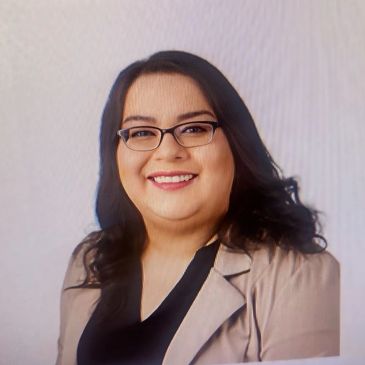 Smiling woman with glasses and dark hair wearing a beige jacket and black top.