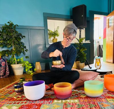 Person playing a singing bowl in a colorful room with plants and cushions.