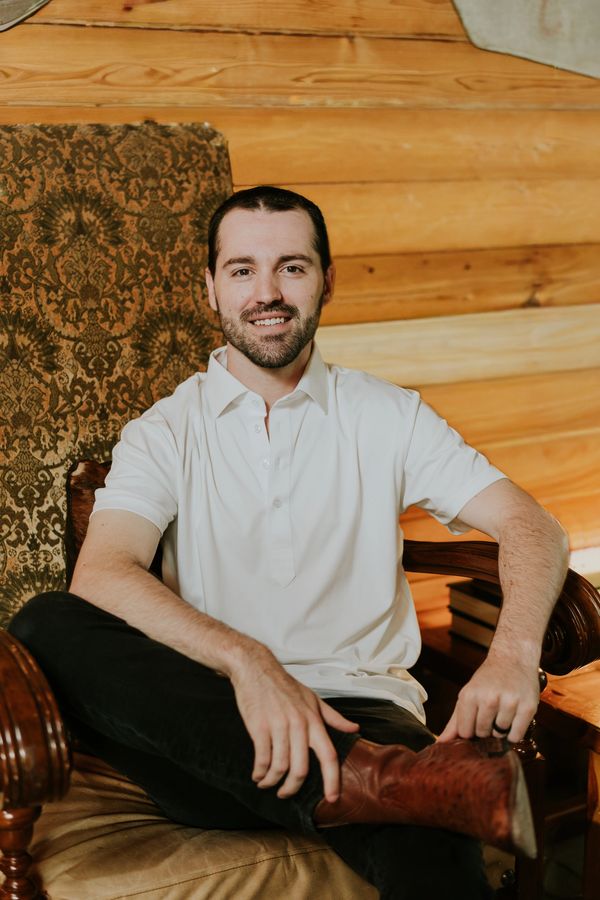 Man in a white polo shirt sitting on a vintage armchair with a wooden wall background.