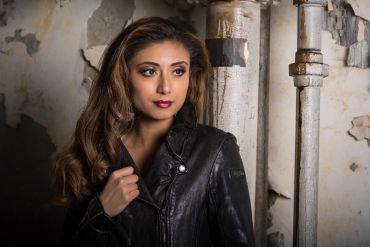 Woman with long hair in a black leather jacket against a rustic wall with pipes.