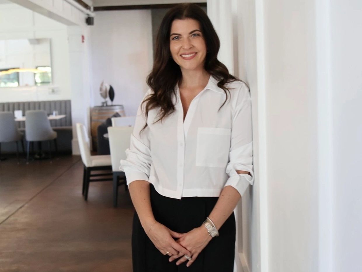 Smiling woman in white blouse and black pants stands indoors near a wall.