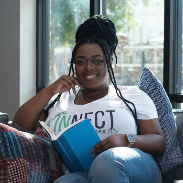 A smiling woman with braided hair reads a blue book, sitting comfortably on a couch.