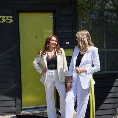 Two women in white suits standing and smiling outside a black building with a yellow door.