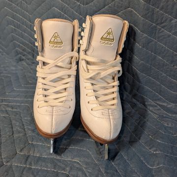 Pair of white figure skates with gold logo on a quilted surface.