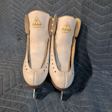 Pair of white figure skates with gold logo on a quilted background.