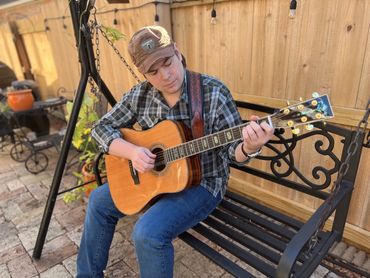 A person playing an acoustic guitar while sitting on a bench outdoors.