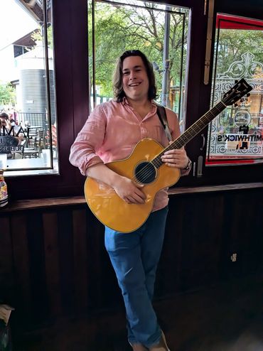 Person smiling and playing an acoustic guitar indoors near windows.