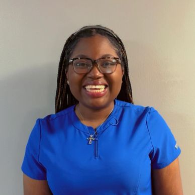 Smiling woman in blue scrubs and glasses against a plain wall.