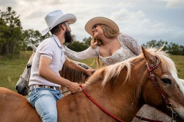 Couple in cowboy hats riding horses and smiling at each other.