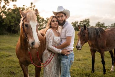 Couple posing with two horses outdoors.