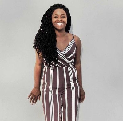 A smiling woman with long braided hair wearing a striped jumpsuit.