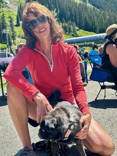 Woman in red jacket smiling with her black pug outdoors on a sunny day.