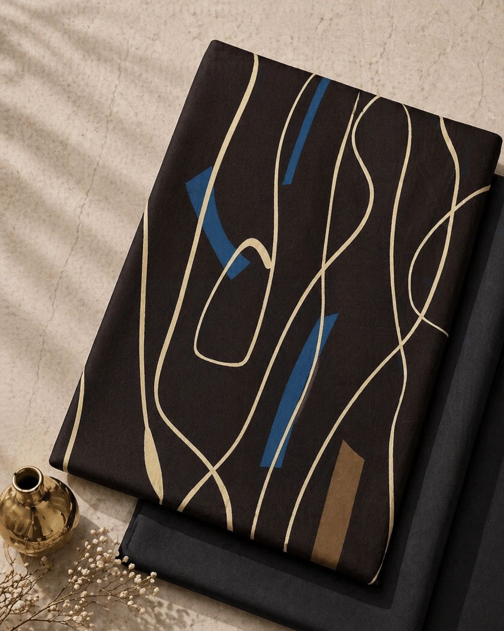 Folded fabric with abstract lines and shapes beside a golden vase and dried flowers.