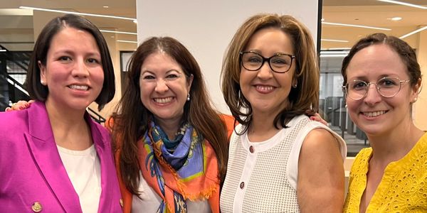 Four women smiling, dressed in bright professional attire, standing close together indoors.