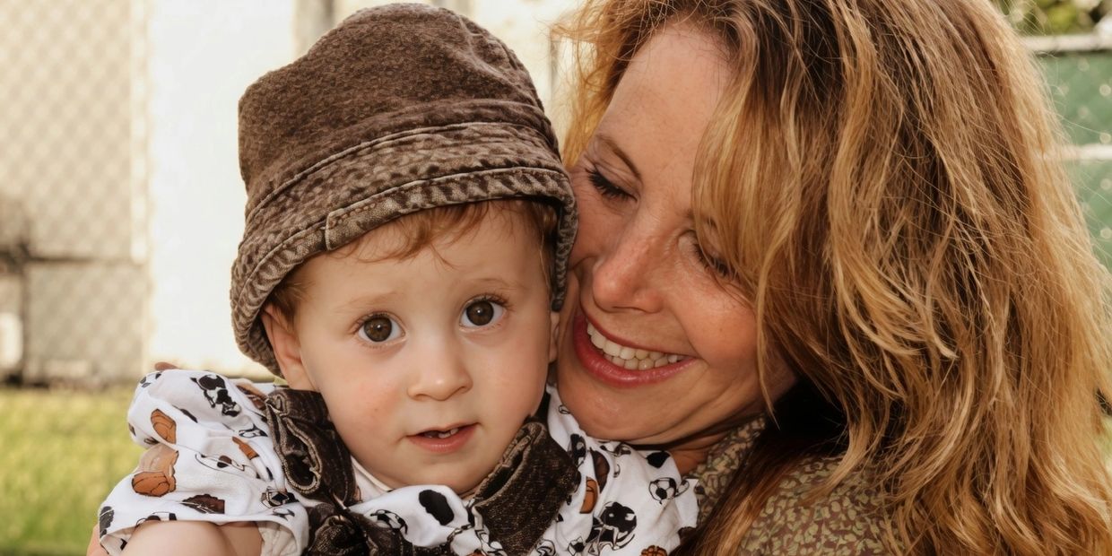 Smiling woman embraces a curious toddler wearing a brown hat outdoors.