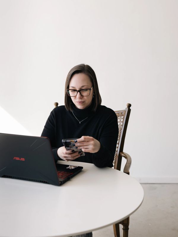 Woman in black sweater using phone at a white round table with a laptop.