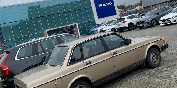 Classic beige Volvo parked in front of a modern Volvo dealership with other cars.