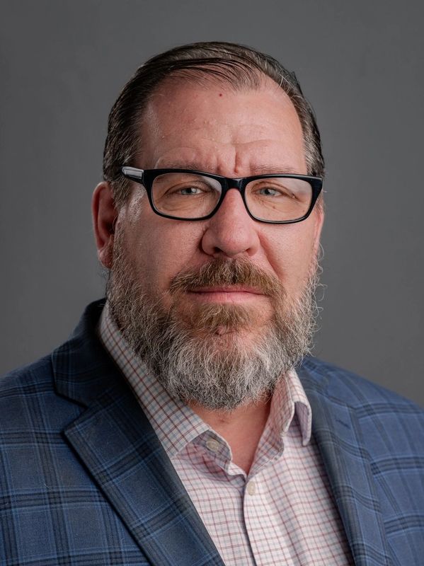 Middle-aged man with glasses, beard, and blue plaid blazer against gray background.